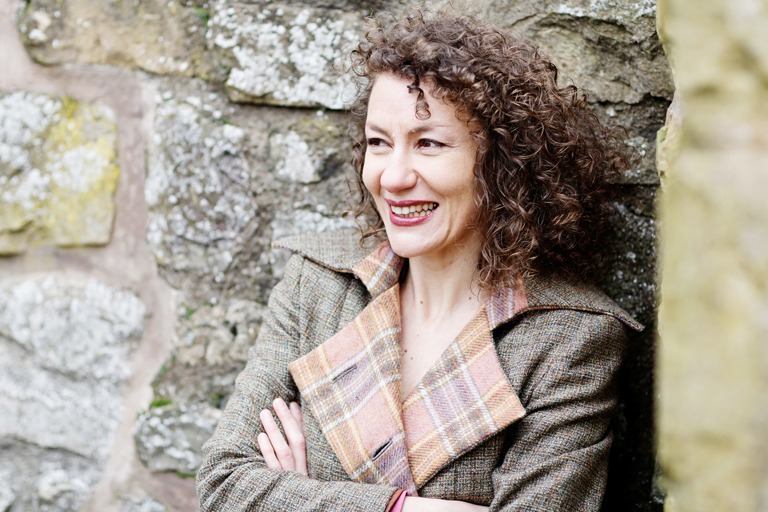 headshot of woman laughing at something out of shot. she is leaning against a brick wall